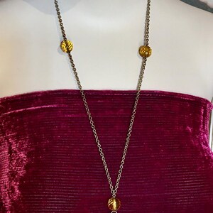 May include: A long gold-toned necklace with amber-colored beads. The necklace features a chain with three decorative amber-colored beads and a tassel of smaller beads. The necklace is displayed on a mannequin wearing a burgundy velvet top.