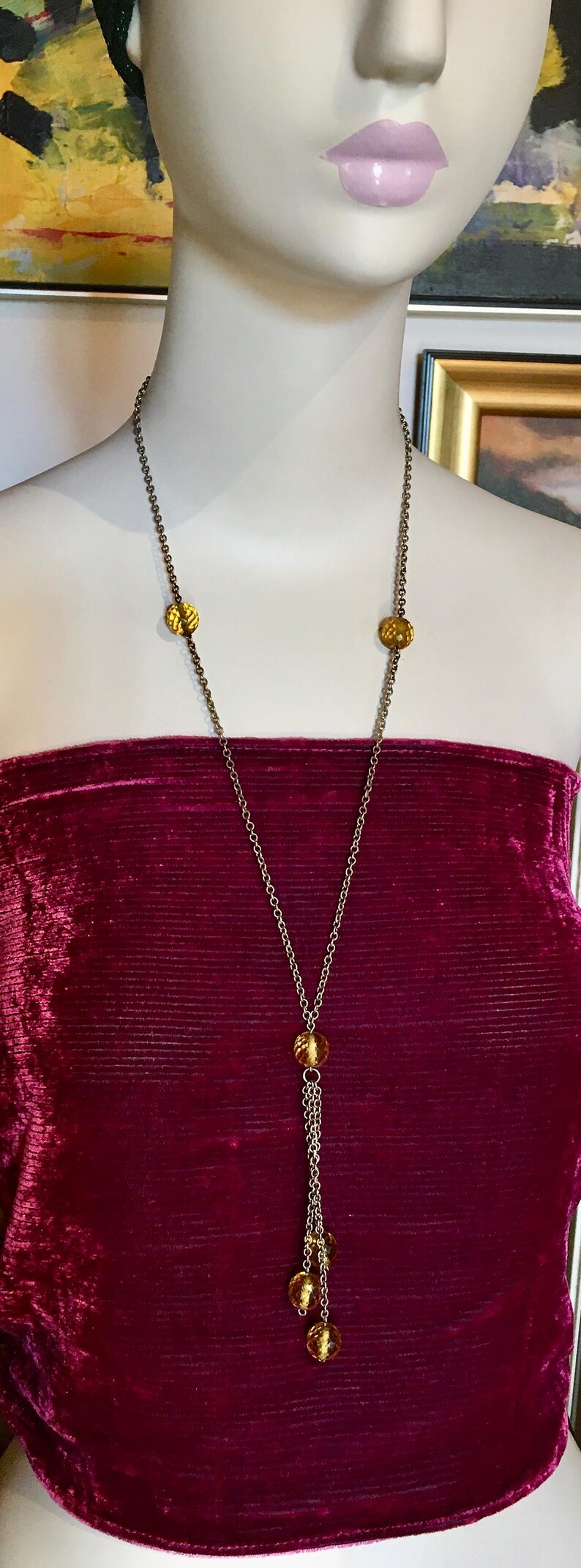 May include: A long gold-toned necklace with amber-colored beads. The necklace features a chain with three decorative amber-colored beads and a tassel of smaller beads. The necklace is displayed on a mannequin wearing a burgundy velvet top.