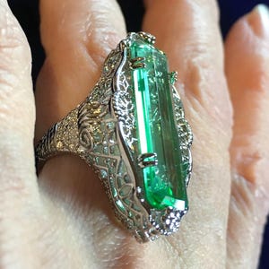 Op de afbeelding: Een opvallende ring met een grote, rechthoekige groene edelsteen. De ring heeft een ingewikkeld filigraanontwerp in een zilveren of witgouden zetting. De smaragdgeslepen steen is het middelpunt, met kleinere accentstenen en gedetailleerde patronen.