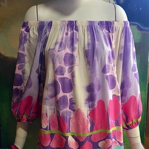 May include: Off-the-shoulder top with a colourful pattern. The top features a mix of purple, pink, and white hues, with a design of irregular shapes and floral elements. The sleeves are three-quarter length, and the hemline has a leopard print.