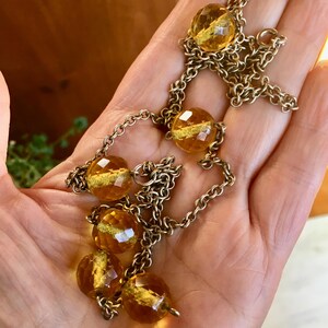 May include: A vintage necklace featuring faceted amber-colored glass beads interspersed with a gold-toned chain. The beads have a golden shimmer within. The necklace is displayed on a hand, showcasing its design and color.