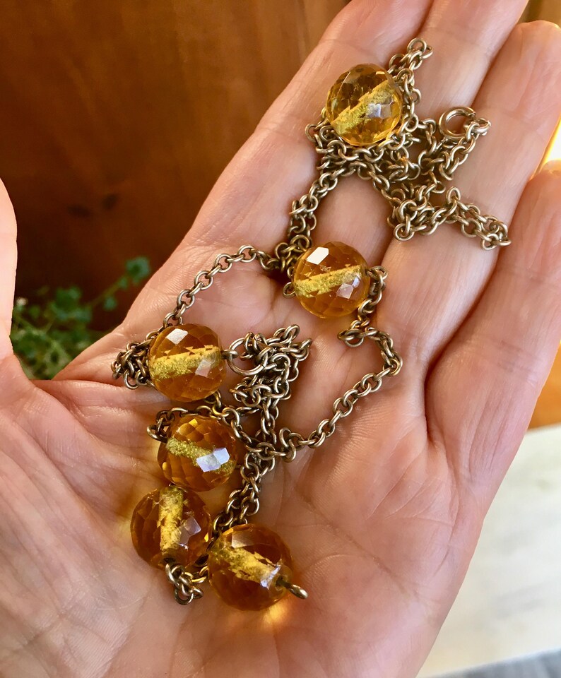 May include: A vintage necklace featuring faceted amber-colored glass beads interspersed with a gold-toned chain. The beads have a golden shimmer within. The necklace is displayed on a hand, showcasing its design and color.