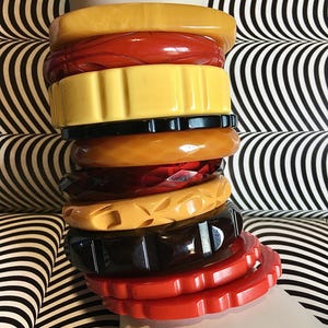 Könnte beinhalten: Ein Stapel aus neun klobigen Armreifen in verschiedenen Farben wie Butterscotch, Rot, Gelb, Schwarz und Bernstein. Die Armbänder weisen unterschiedliche Texturen auf, darunter glatte Oberflächen, geometrische Schnitzmuster und gekerbte Ränder.