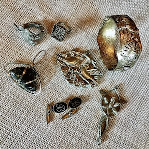 Może przedstawiać: Asortyment biżuterii vintage, w tym srebrna bransoletka z kwiatowymi detalami, pierścionki, wisiorek, spinki do mankietów z literą "B" i broszka w kształcie ptaka. Przedmioty ułożone są na teksturowanej, neutralnej powierzchni.