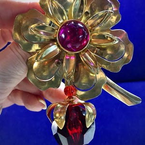 May include: A gold-toned flower brooch with a large red rhinestone in the center. The flower has a dangling red crystal drop with gold accents.