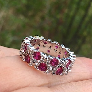 May include: A silver-toned band ring featuring a repeating pattern of round, vibrant pink stones set within a metallic frame. The design includes small, clear accent stones interspersed between the larger pink circular gems along the entire band.