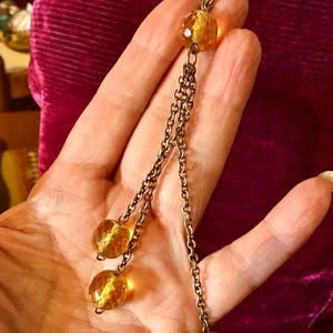 May include: A vintage-style necklace with a long chain and three dangling strands. Each strand features a faceted, amber-colored bead. The necklace has a bronze-toned chain and beads, creating a classic, elegant look.
