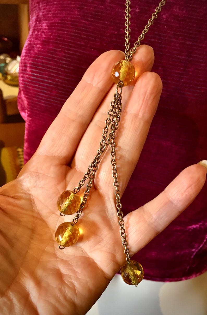 May include: A vintage-style necklace with a long chain and three dangling strands. Each strand features a faceted, amber-colored bead. The necklace has a bronze-toned chain and beads, creating a classic, elegant look.