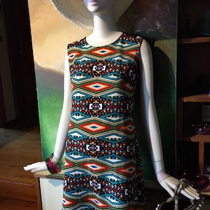 Op de afbeelding: Een mouwloze jurk met een kleurrijk geometrisch patroon in rood, blauw, groen en geel wordt getoond op een mannequin. De mannequin draagt een breedgerande hoed, een rode zonnebril en armbanden. Een strooien tas met witte accenten staat ernaast.
