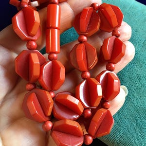 May include: A vintage red necklace featuring geometric, faceted beads and small round beads. The necklace is held in a hand, showcasing the rich color and intricate design. The beads are strung together, creating a bold statement piece.