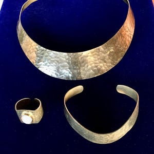 Może przedstawiać: Zestaw trzech złotych, młotkowanych ozdób metalowych: szeroki naszyjnik choker, bransoletka i pierścionek z białym kamieniem.