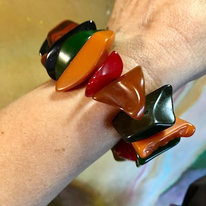 Könnte beinhalten: Ein farbenfrohes Armband mit gebogenen, dreieckigen Perlen in Rot-, Orange-, Grün- und Brauntönen. Das Armband, das am Handgelenk getragen wird, besteht aus glänzendem Material. Die Perlen sind etwa 2,5 cm lang.