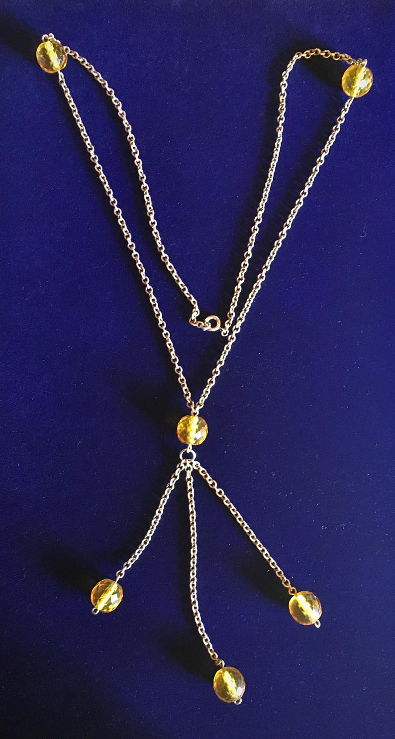 May include: A gold-colored necklace with a delicate chain design. The necklace features multiple strands and is adorned with yellow, faceted beads. The beads are evenly spaced along the chain, creating a symmetrical and elegant look. The necklace is displayed against a dark blue background.