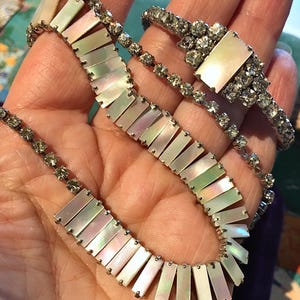 Può includere: Un set di gioielli vintage con collana e bracciale. La collana e il bracciale sono realizzati con segmenti rettangolari iridescenti di madreperla, collegati da metallo argentato. Il bracciale ha un elemento centrale rettangolare in madreperla circondato da strass trasparenti.