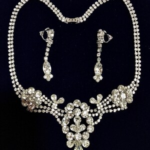 May include: A silver-toned necklace and earring set featuring clear, faceted rhinestones. The necklace has a double-strand chain leading to a central cluster of round and marquise-cut stones. The matching drop earrings feature a similar design.