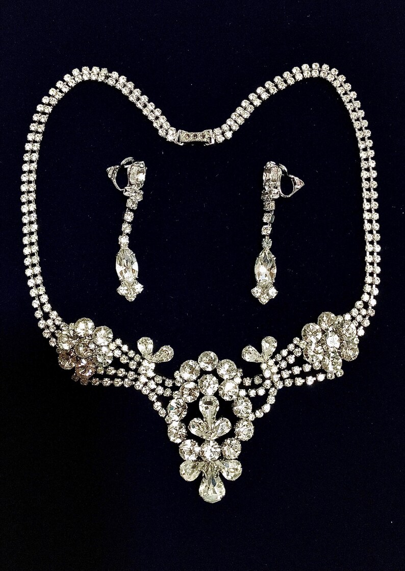 May include: A silver-toned necklace and earring set featuring clear, faceted rhinestones. The necklace has a double-strand chain leading to a central cluster of round and marquise-cut stones. The matching drop earrings feature a similar design.