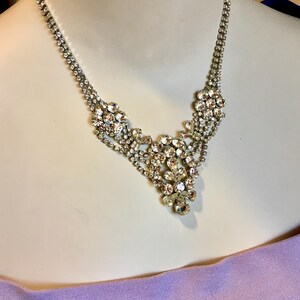 May include: A silver-toned necklace featuring a central cluster of clear, faceted rhinestones arranged in a floral-inspired pattern. The chain consists of two delicate rows of small, round rhinestones that meet at the ornate, multi-layered centerpiece.