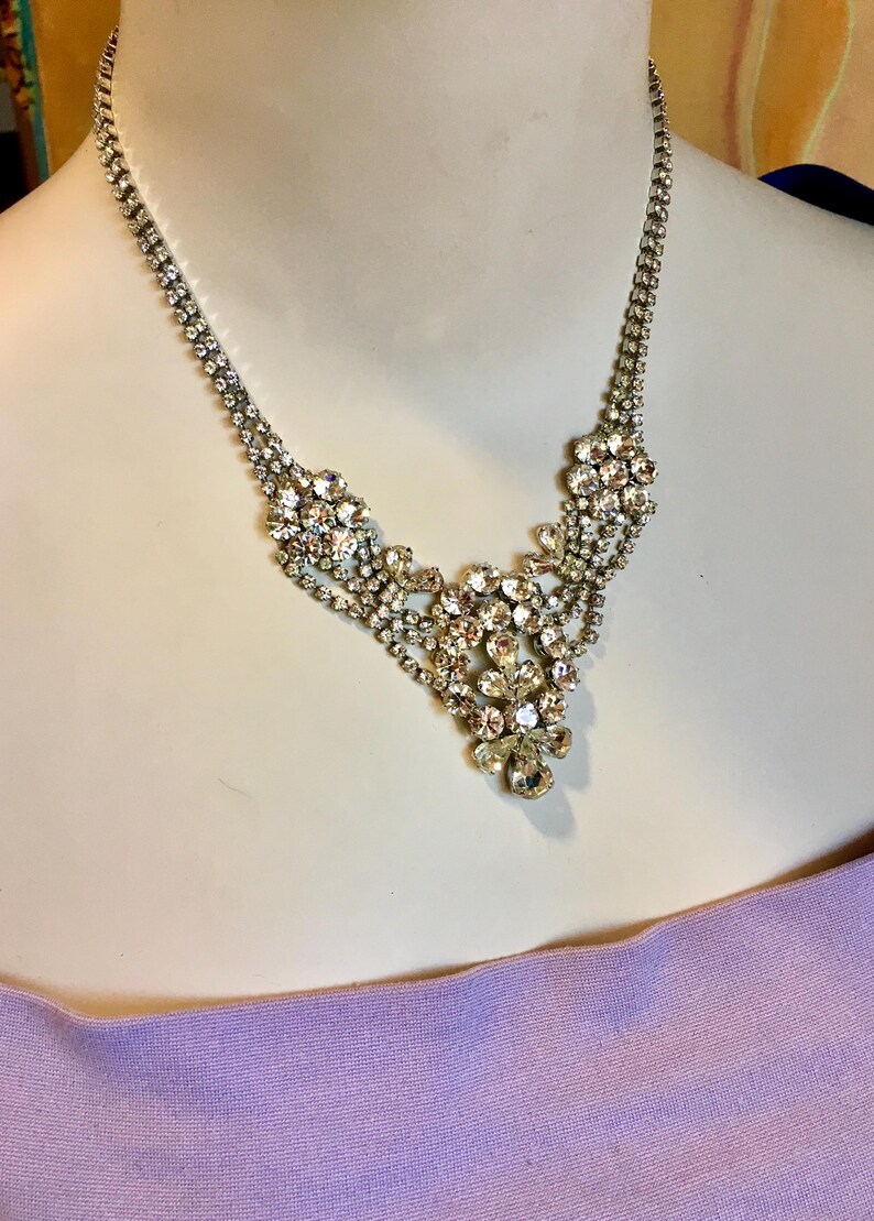 May include: A silver-toned necklace featuring a central cluster of clear, faceted rhinestones arranged in a floral-inspired pattern. The chain consists of two delicate rows of small, round rhinestones that meet at the ornate, multi-layered centerpiece.