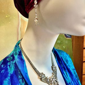 May include: A silver-toned necklace and matching drop earring set featuring clear, faceted rhinestones. The necklace has a multi-strand design that converges into a central cluster of stones, while the earring features a vertical arrangement.