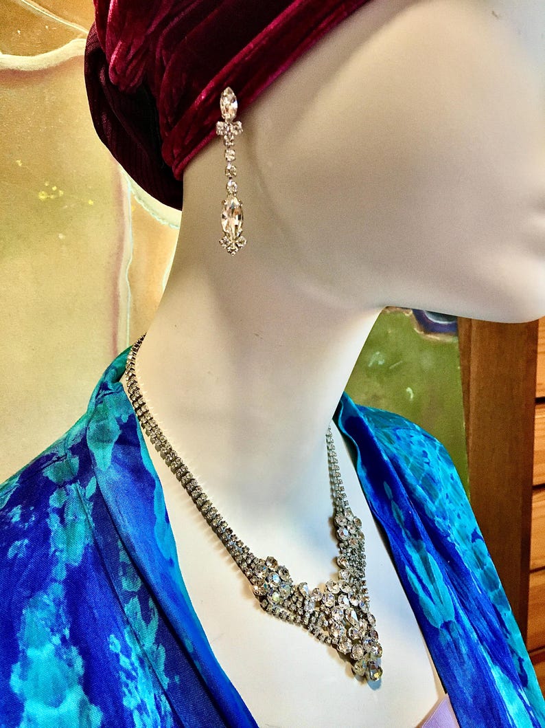 May include: A silver-toned necklace and matching drop earring set featuring clear, faceted rhinestones. The necklace has a multi-strand design that converges into a central cluster of stones, while the earring features a vertical arrangement.