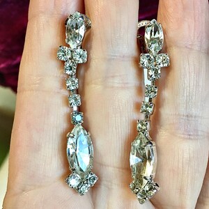 May include: A pair of silver-toned drop earrings featuring a vertical arrangement of clear, faceted rhinestones. Each earring includes a marquise-cut stone at the base, a series of smaller square-cut stones, and a top marquise-cut stone.