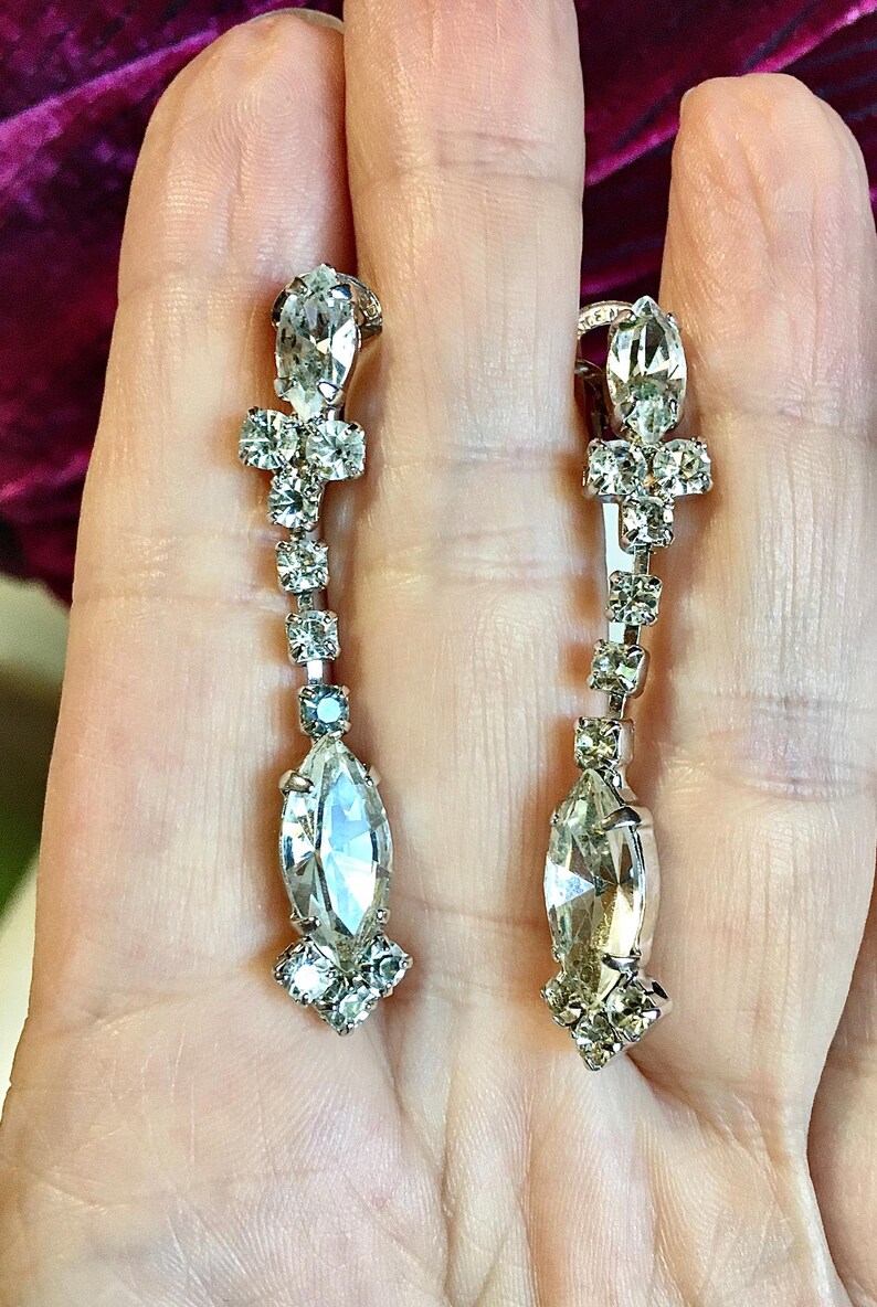 May include: A pair of silver-toned drop earrings featuring a vertical arrangement of clear, faceted rhinestones. Each earring includes a marquise-cut stone at the base, a series of smaller square-cut stones, and a top marquise-cut stone.