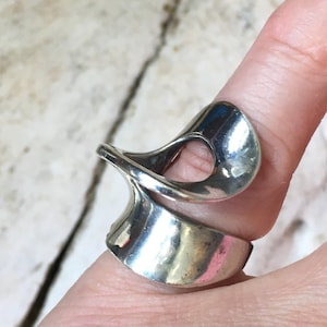 Op de afbeelding: Een gepolijste zilverkleurige ring met een brede, gladde band die naar boven toe draait in een open, sculpturaal lusontwerp. Het metalen oppervlak is reflecterend en gebogen, wat een continue, vloeiende vorm rond de vinger creëert.