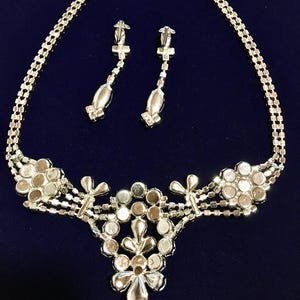 May include: A silver-toned jewelry set consisting of a necklace and a pair of drop earrings. The necklace features a central floral-inspired cluster of round clear rhinestones, with additional floral accents and a multi-strand chain design.
