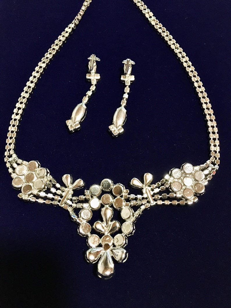 May include: A silver-toned jewelry set consisting of a necklace and a pair of drop earrings. The necklace features a central floral-inspired cluster of round clear rhinestones, with additional floral accents and a multi-strand chain design.