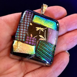 Pode incluir: Um pingente de vidro preto com um design de libélula e detalhes iridescentes em verde, roxo e dourado.