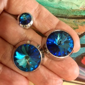Op de afbeelding: Drie sieraden met levendige blauwe kristallen stenen. Twee zijn groot, rond en gezet in zilverkleurig metaal, waarschijnlijk manchetknopen. De derde is kleiner, mogelijk een oorbel. De kristallen hebben een facetontwerp.
