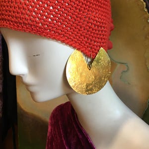 Könnte beinhalten: Eine leuchtend orangefarbene Häkelmütze auf einem weißen Schaufensterpuppenkopf, kombiniert mit einem großen, runden, strukturierten Ohrring aus goldfarbenem Metall mit keilförmigem Ausschnitt. Die Puppe ist mit tief magenta-farbenem Samtstoff drapiert.