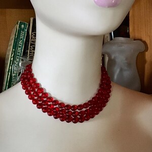 May include: A three-strand red beaded necklace. The beads are small and round, and the necklace is displayed on a mannequin. The necklace is a vibrant shade of red, and the beads appear to be made of glass or a similar material.