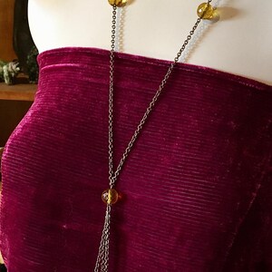 May include: A long, antique-style necklace with a chain and amber-colored beads. The necklace is displayed on a mannequin wearing a burgundy velvet top. The necklace has a unique, layered design.