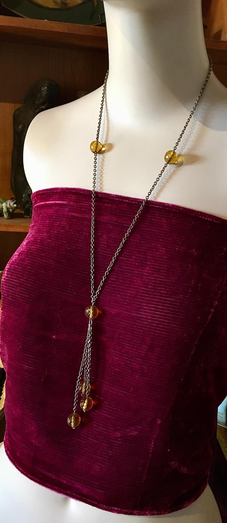May include: A long, antique-style necklace with a chain and amber-colored beads. The necklace is displayed on a mannequin wearing a burgundy velvet top. The necklace has a unique, layered design.