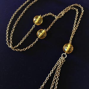 May include: A gold-toned necklace with multiple strands and amber-colored, faceted beads. The necklace features a long chain with three dangling strands and several round beads. The beads are a translucent yellow color.