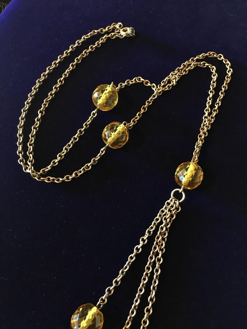 May include: A gold-toned necklace with multiple strands and amber-colored, faceted beads. The necklace features a long chain with three dangling strands and several round beads. The beads are a translucent yellow color.