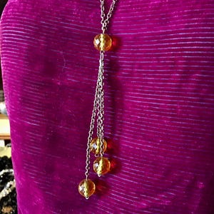 May include: A long, silver-toned necklace with a series of amber-colored, faceted beads. The necklace has a central bead and a cascading design with three additional beads. The background is a deep purple fabric.