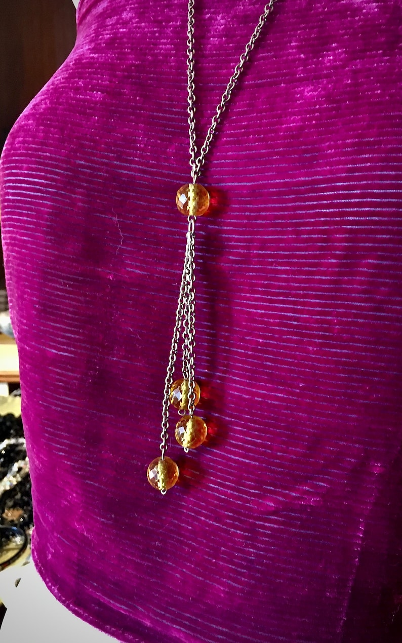 May include: A long, silver-toned necklace with a series of amber-colored, faceted beads. The necklace has a central bead and a cascading design with three additional beads. The background is a deep purple fabric.