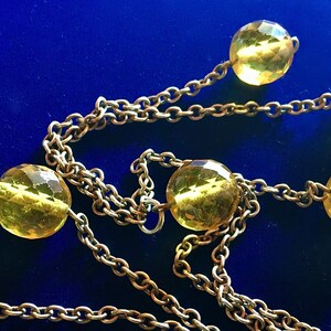 May include: A gold-toned necklace with faceted yellow beads and a delicate chain. The beads are evenly spaced along the chain, creating a simple yet elegant design. The necklace is set against a deep blue background.