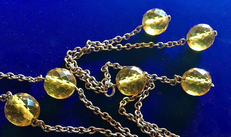 May include: A gold-toned necklace with faceted yellow beads and a delicate chain. The beads are evenly spaced along the chain, creating a simple yet elegant design. The necklace is set against a deep blue background.