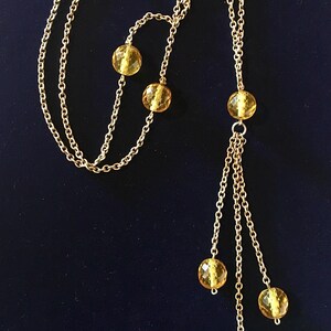May include: A gold-toned necklace with a delicate chain design. The necklace features several amber-colored, faceted beads. The beads are interspersed along the chain and also dangle in a cluster. The necklace is displayed against a dark blue background.