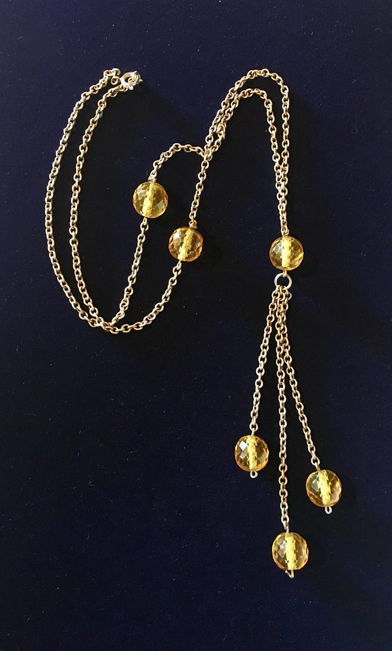 May include: A gold-toned necklace with a delicate chain design. The necklace features several amber-colored, faceted beads. The beads are interspersed along the chain and also dangle in a cluster. The necklace is displayed against a dark blue background.
