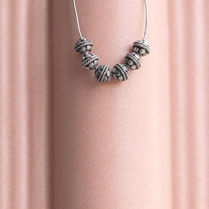 May include: A silver necklace featuring seven textured, spherical beads strung on a thin, silver wire. The beads have a detailed, patterned design. The background is a soft, pinkish-peach color.
