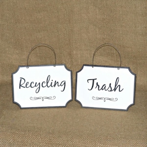 May include: Two white signs with black borders and black lettering. The signs are hanging from a wire and say "Recycling" and "Trash".