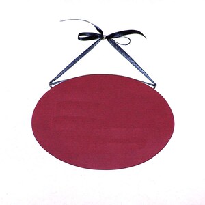 May include: A blank, oval-shaped sign with a burgundy red background. The sign is hanging from a black ribbon.