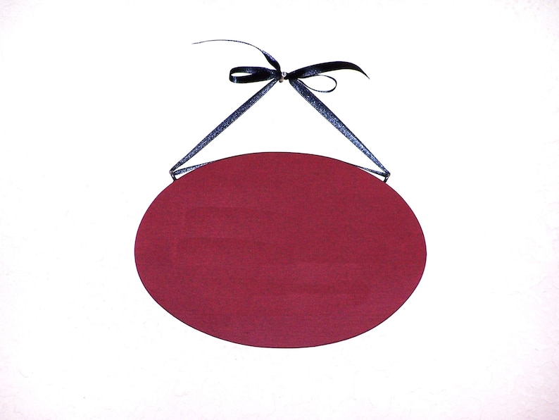 May include: A blank, oval-shaped sign with a burgundy red background. The sign is hanging from a black ribbon.