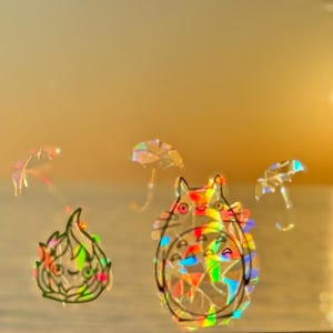 May include: Holographic stickers featuring cartoon characters and umbrellas. The stickers have a rainbow effect, with a cartoon cat-like character and a flame-like character. The background is a blurred view of the ocean and sky at sunset.