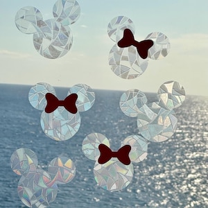 May include: Decorative window clings in the shape of Mickey Mouse heads. The clings have a mosaic-like design with iridescent, shimmering details. Some clings feature a dark red bow. The background shows a view of the ocean and sky.