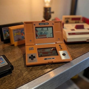 K&ouml;nnte beinhalten: Orangefarbene Nintendo Game & Watch Handheld-Konsole mit dem Spiel Donkey Kong. Die Konsole hat einen kreuzf&ouml;rmigen Controller und eine Starttaste. Mehrere Spielkassetten und eine klassische Nintendo-Konsole sind im Hintergrund zu sehen.